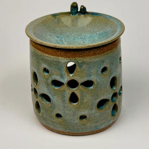 May include: A ceramic teal and brown jar with a lid. The jar has a cylindrical shape with decorative cut-out patterns. The lid has a small handle. The jar is glazed with a mottled teal and brown finish.