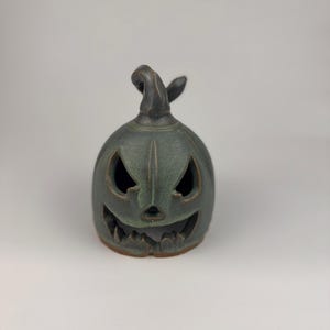 May include: A dark green ceramic jack-o'-lantern candle holder with a carved, jagged face featuring triangular eyes, a small nose, and a wide, toothy grin. The pumpkin-shaped object has a curved stem on top and a smooth, matte finish.