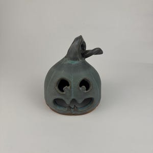 May include: A dark gray ceramic jack-o'-lantern figurine with a smooth, matte finish. The pumpkin-shaped object features two circular eye cutouts and a wide, jagged mouth opening, topped with a curved, twisted stem extending to the right.