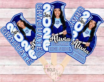 May include: Graduation photo props featuring a graduate in a blue cap and gown. The props are light blue and white with the text "Class of 2026 GRADUATE" and the graduate's name and high school. The props are on wooden sticks.