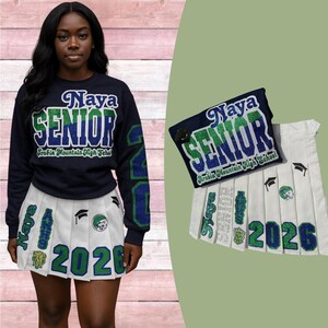 May include: Navy blue sweatshirt and white pleated skirt with green and blue accents. The sweatshirt says "Naya SENIOR Arabia Mountain High School" and the skirt says "2026" with graduation-themed patches. A matching patch is also shown.
