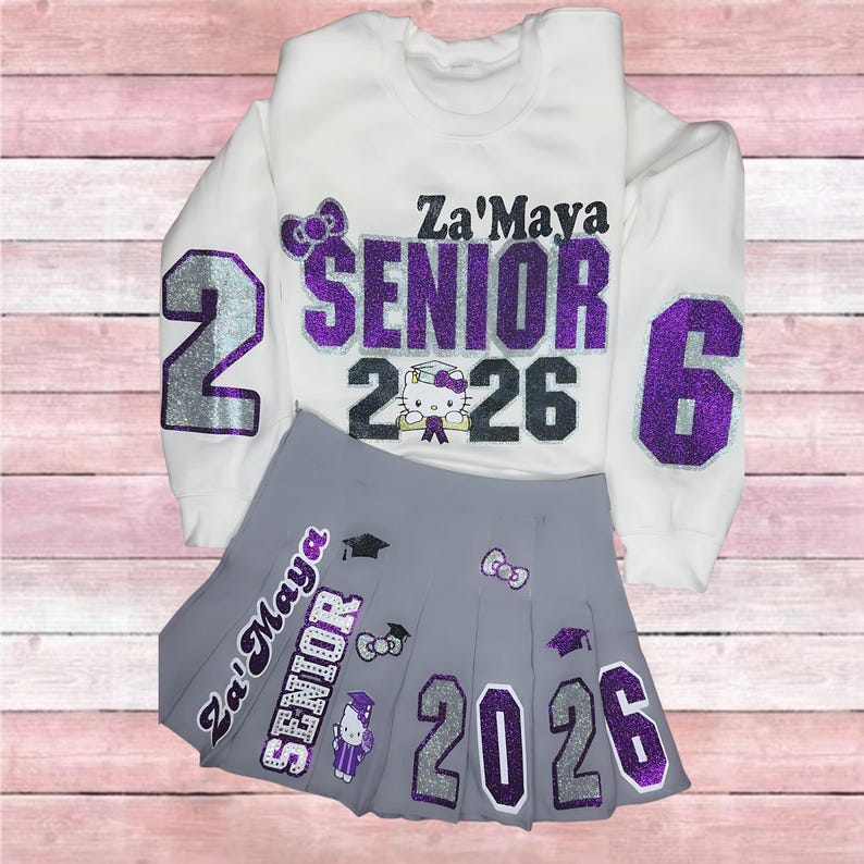 Może przedstawiać: Biała bluza i szara plisowana sp&oacute;dnica. Bluza ma napis "Za'Maya SENIOR 2026" w fioletowym i srebrnym brokacie. Sp&oacute;dnica ma pasujący tekst i grafikę Hello Kitty. Str&oacute;j z motywem ukończenia szkoły.