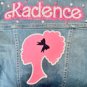 May include: A light blue denim jacket with a pink and white appliqué of the word "Kadence" and a pink silhouette of a woman with a black bow in her hair.