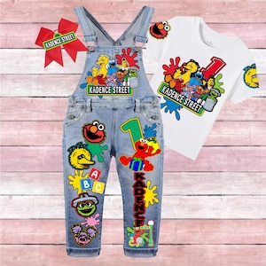 May include: A light blue denim overall set with a white t-shirt and a red hair bow. The clothing features colorful Sesame Street character graphics, the text Kadence Street, the number 1, and ABC blocks in a vibrant, multi-colored design.