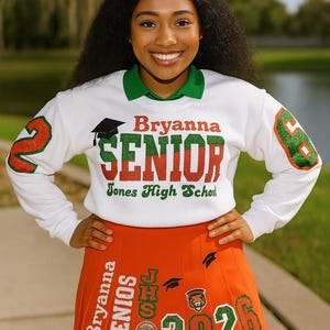 May include: A white sweatshirt with green collar and sleeves, featuring the name "Bryanna" and the word "SENIOR" in red and green glitter lettering. An orange pleated skirt with "2026" and "SENIORS" is also visible. The image is taken outdoors.