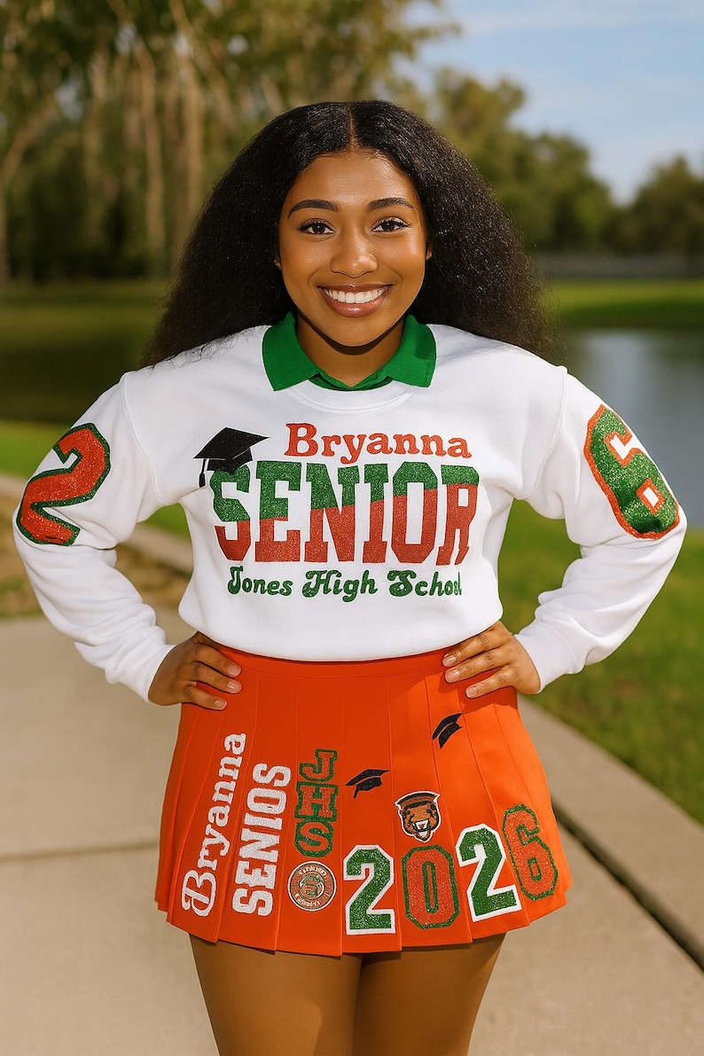 May include: A white sweatshirt with green collar and sleeves, featuring the name "Bryanna" and the word "SENIOR" in red and green glitter lettering. An orange pleated skirt with "2026" and "SENIORS" is also visible. The image is taken outdoors.