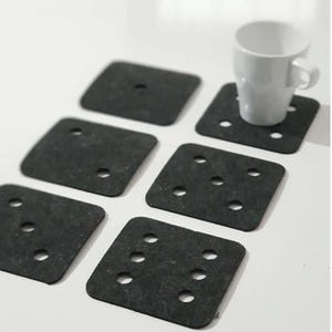Könnte beinhalten: Ein Set aus sechs dunkelgrauen quadratischen Untersetzern mit abgerundeten Ecken und kleinen kreisförmigen Aussparungen. Eine weiße Keramik-Tasse steht auf einem Untersetzer. Die Untersetzer sind auf einer weißen Oberfläche angeordnet.