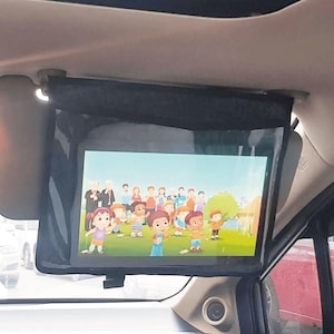 May include: A black, rectangular car entertainment system is mounted to the ceiling. The screen displays a cartoon with children. The system is installed in a car, with the dashboard and a portion of the sunroof visible.