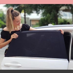 Peut inclure: Une femme tient un pare-soleil de fenêtre de voiture noir. Le pare-soleil est rectangulaire et fabriqué en matériau maillé. La voiture est blanche, et l'arrière-plan est flou avec des arbres et un bâtiment.