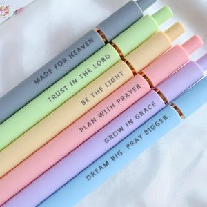 Puede incluir: Un conjunto de bolígrafos de colores pastel con detalles dorados. Cada bolígrafo lleva una inscripción blanca con una frase inspiradora, como "MADE FOR HEAVEN" y "DREAM BIG. PRAY BIGGER."