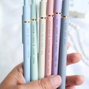 Hand holding six pastel Catholic engraved pens in blue, mint green, cream, pink, lavender, and gray, each with inspirational Bible verse engravings such as 'Jesus, I Trust in You' and 'Love Like Jesus'.