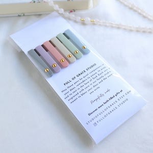 Back view of Holy Reminders Catholic Pen Set packaging with Full of Grace Studio message card and pastel pens showing gold clip accents, styled with rosary beads and journal.