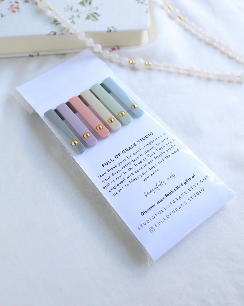 Back view of Holy Reminders Catholic Pen Set packaging with Full of Grace Studio message card and pastel pens showing gold clip accents, styled with rosary beads and journal.
