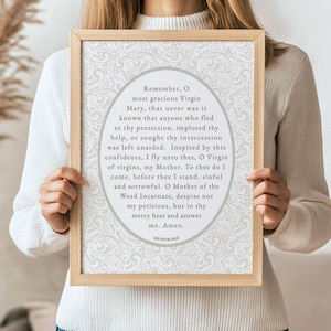 Woman holding a framed Memorare Prayer printable wall art with beige floral background — minimalist Catholic digital download artwork featuring Marian devotion for faith-filled homes.