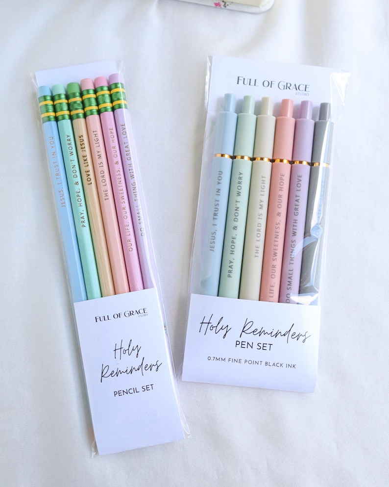 Holy Reminders Catholic Pencil Set and Pen Set from Full of Grace Studio displayed side by side in clear packaging, featuring pastel colors and engraved inspirational messages.
