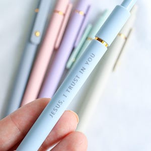 Close-up of pastel blue Catholic engraved gel pen reading 'Jesus, I Trust in You,' with other pastel faith-inspired pens blurred in the background.