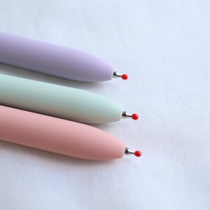 Close-up of three pastel gel pen tips in lavender, mint green, and pink showing fine point black ink tips with protective red covers, on white fabric background.