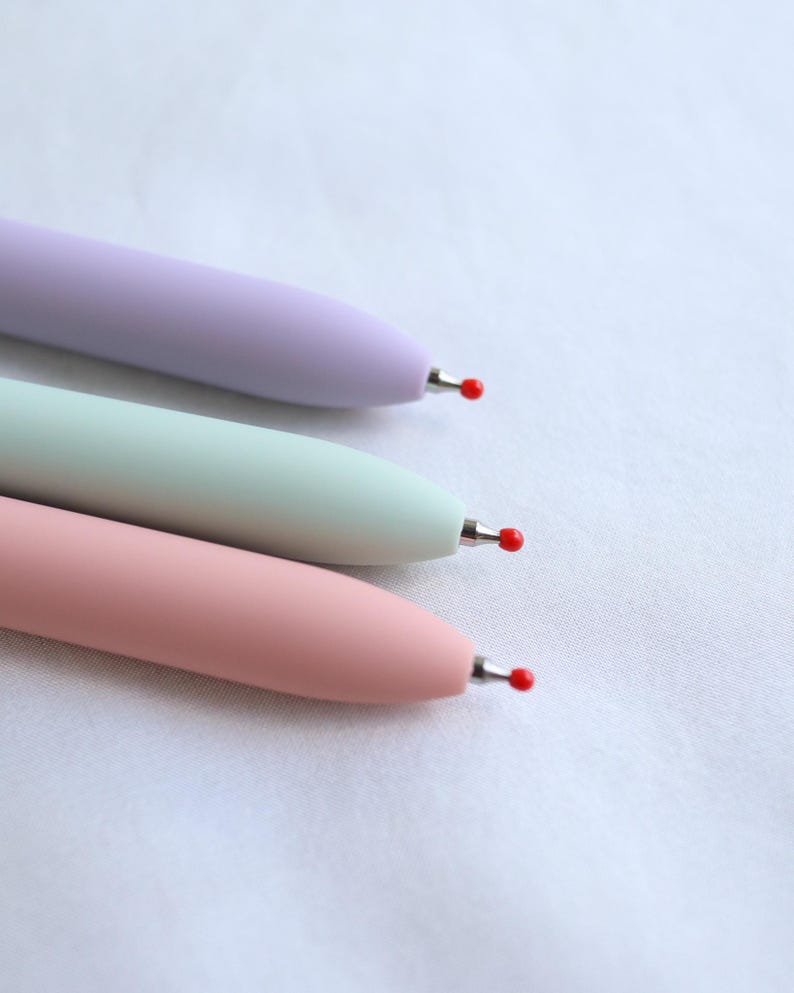 Close-up of three pastel gel pen tips in lavender, mint green, and pink showing fine point black ink tips with protective red covers, on white fabric background.