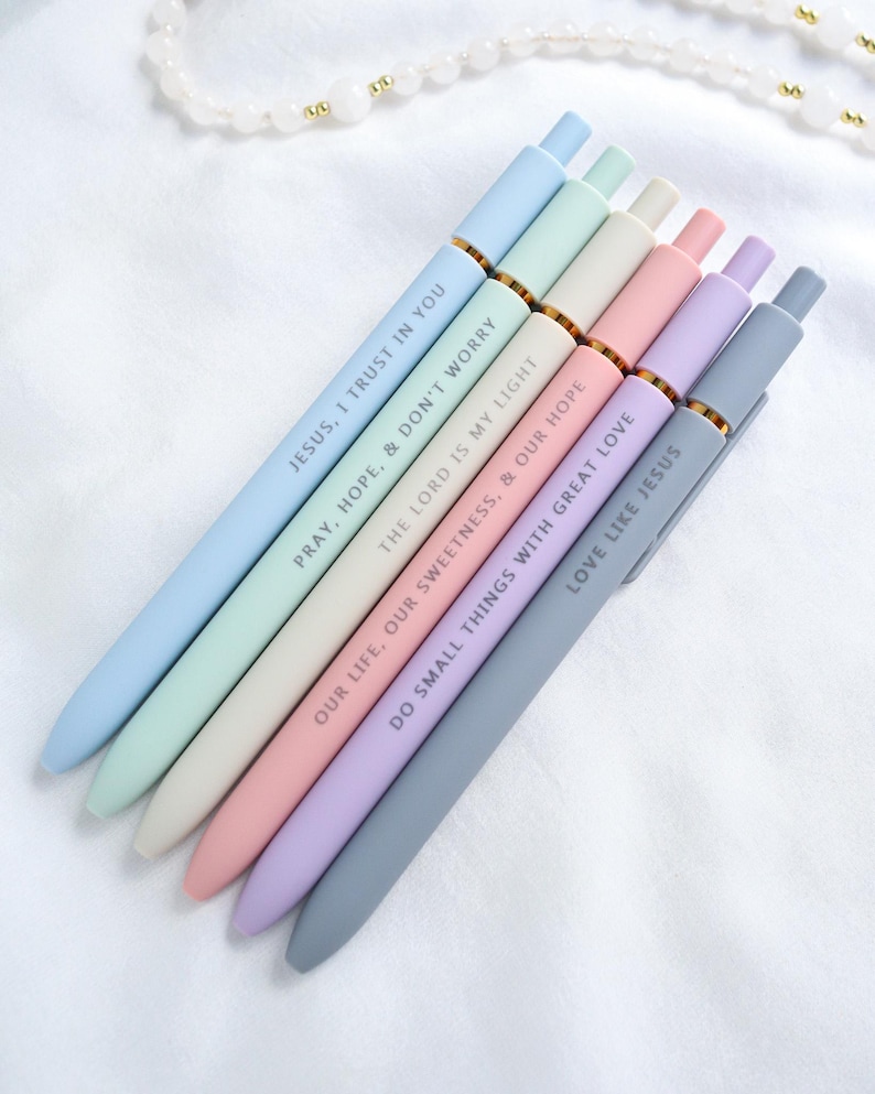 Set of six pastel Catholic engraved gel pens in blue, green, cream, pink, lavender, and gray with inspirational Bible verse messages, displayed on a white cloth with rosary beads.