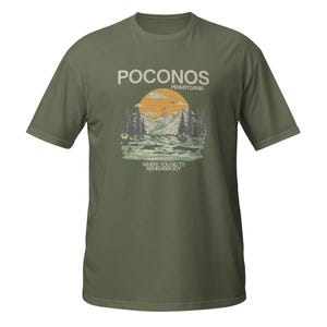 Op de afbeelding: Een olijfgroen t-shirt met korte mouwen en een grafische print van een berglandschap met een grote oranje zon, dennenbomen en vogels. De tekst luidt POCONOS PENNSYLVANIA en WHERE YOU GO TO REMEMBER JOY in een crèmekleurig lettertype.