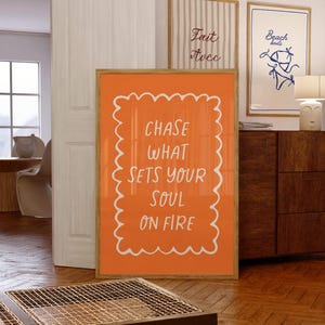 Op de afbeelding: Een oranje ingelijste print met de witte tekst "CHASE WHAT SETS YOUR SOUL ON FIRE". Twee andere ingelijste prints zijn zichtbaar, één met verticale strepen en de andere met een blauwe bikini-illustratie. De prints worden tentoongesteld in een kamer met houten vloeren.