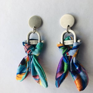 May include: A pair of silver earrings with a knotted fabric design. The fabric is a mix of blue, green, purple, and orange colors. The earrings have a circular silver top.