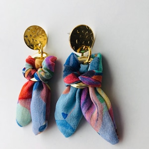 以下が含まれることがあります： 金色の金属製のアクセントが付いた、カラフルな布製のイヤリングのペア。布は青、ピンク、緑のパターンです。金色の金属はハンマードテクスチャです。