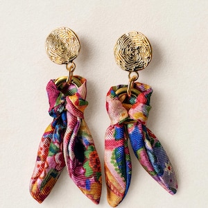 May include: A pair of dangle earrings featuring circular gold-tone metal studs with a textured, swirling pattern. Each stud connects to a gold-tone ring holding a knotted piece of multi-colored fabric with a vibrant floral and abstract print.
