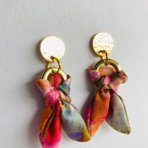 Puede incluir: Un par de pendientes de oro con un lazo de tela colorido. La tela es una mezcla de rosa, naranja, amarillo y azul.