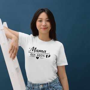 Op de afbeelding: Wit T-shirt met het woord "Mama" in scriptlettertype, "EST. 2026", een hartje en babyvoetjes. Het shirt wordt gedragen door een persoon die leunt op een witte ladder tegen een donkerblauwe achtergrond.
