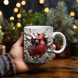 May include: White ceramic mug with a striking red cardinal bird design. The bird is depicted emerging from a textured, off-white surface. The mug has a white handle. The background is blurred.