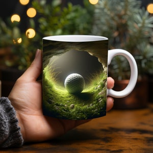 May include: White ceramic mug with a golf ball resting in a green grassy area inside a dark cave. The mug has a white handle.