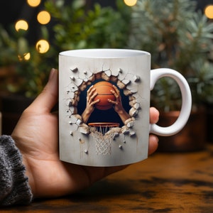 Puede incluir: Taza de cerámica blanca con un diseño de baloncesto. Una mano rompe una pared agrietada sosteniendo una pelota de baloncesto y se ve un aro de baloncesto.