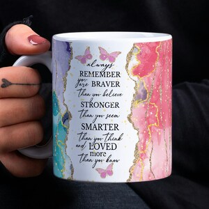 May include: White ceramic mug with a pink and blue watercolor design and gold glitter. The mug has a quote that reads "always remember you are braver than you believe stronger than you seem smarter than you think and loved more than you know".