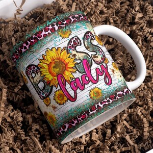 May include: White ceramic mug with a pink, black, and turquoise design. The mug has a rustic woodgrain background with the words "Boss Lady" in pink lettering. The design also features sunflowers and a leopard print pattern.