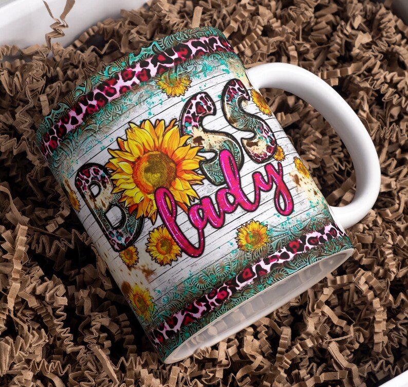 May include: White ceramic mug with a pink, black, and turquoise design. The mug has a rustic woodgrain background with the words "Boss Lady" in pink lettering. The design also features sunflowers and a leopard print pattern.