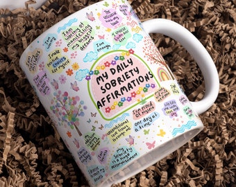 May include: White ceramic mug with a colourful design featuring butterflies, flowers, and the text "My Daily Sobriety Affirmations". The mug has a handle and is filled with positive affirmations.