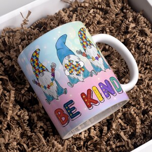 Puede incluir: Una taza de cer&aacute;mica blanca con un dise&ntilde;o colorido que presenta cuatro gnomos con piezas de rompecabezas y el texto "Be Kind".