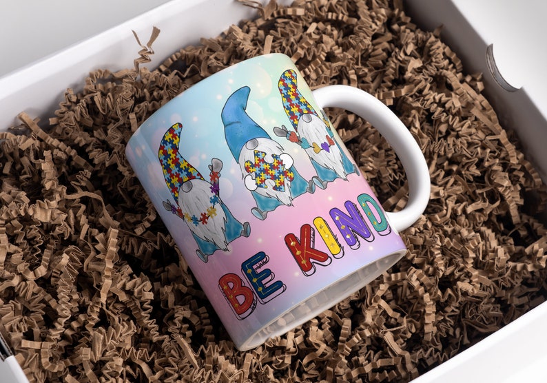 Puede incluir: Una taza de cer&aacute;mica blanca con un dise&ntilde;o colorido que presenta cuatro gnomos con piezas de rompecabezas y el texto "Be Kind".