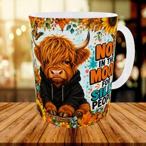 May include: A white ceramic mug featuring a graphic of a fluffy brown Highland cow wearing a black hoodie, surrounded by autumn leaves, sunflowers, and a butterfly. The text reads NOT IN THE MOOD FOR SILLY PEOPLE in orange and teal block letters.