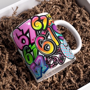 Könnte beinhalten: Weiße Keramiktasse mit einem farbenfrohen Graffiti-Design mit den Zahlen 6 und 7 in verschiedenen Farben. Die Tasse hat einen weißen Henkel und befindet sich in einer Box mit braunem Verpackungsmaterial.