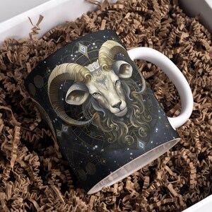 May include: A white ceramic mug with a black, gold, and white design of a ram's head with a starry background.