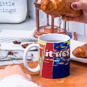 May include: White ceramic mug with a colorful design featuring the text "Titties" and a variety of candy-like shapes. The mug is filled with a brown liquid and is sitting on a wooden surface. A hand is holding a croissant over the mug.
