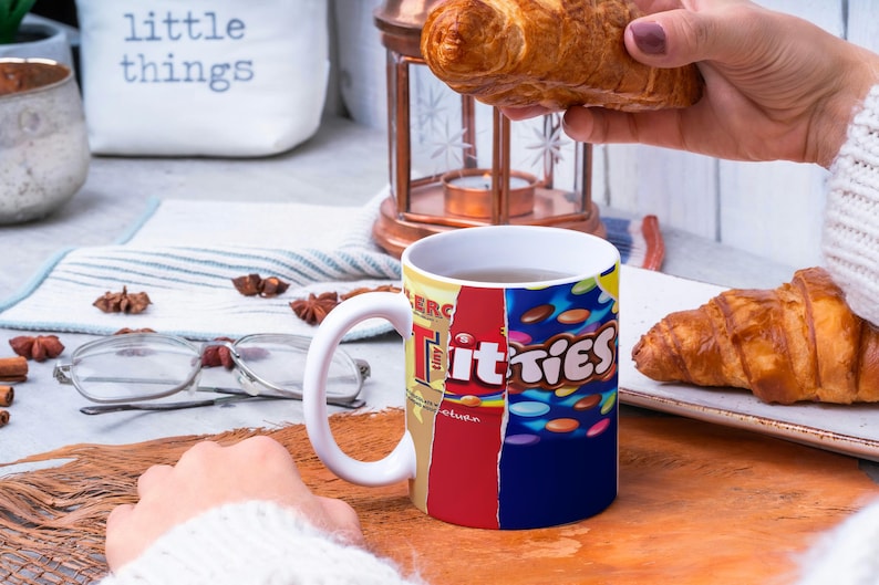 May include: White ceramic mug with a colorful design featuring the text "Titties" and a variety of candy-like shapes. The mug is filled with a brown liquid and is sitting on a wooden surface. A hand is holding a croissant over the mug.