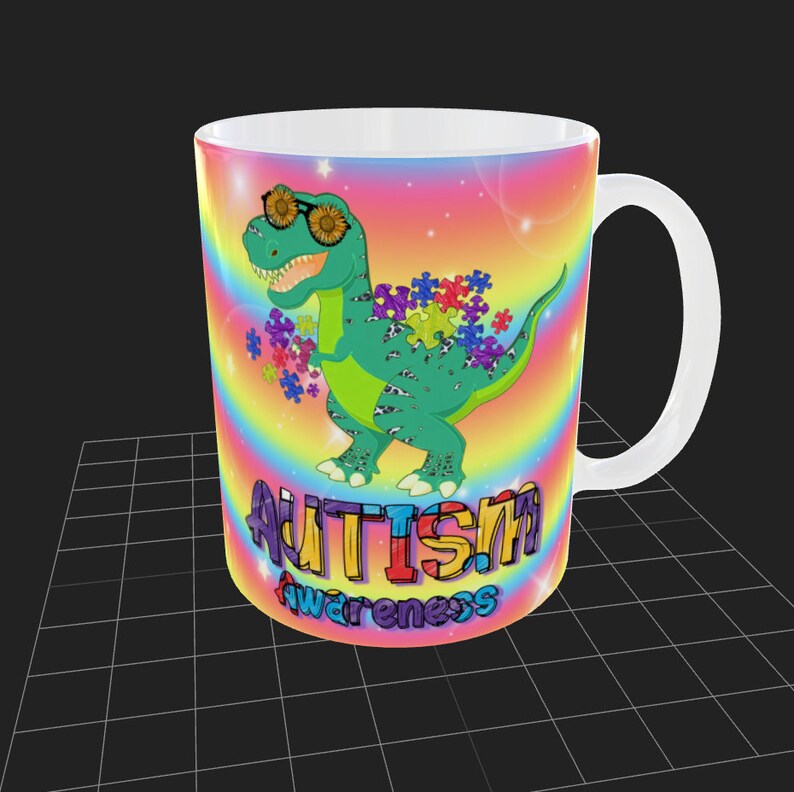 Puede incluir: Una taza de cer&aacute;mica blanca con un fondo de arco&iacute;ris colorido y un dinosaurio verde con gafas de sol. El dinosaurio sostiene piezas de rompecabezas y el texto "Autism Awareness" est&aacute; impreso debajo del dinosaurio.