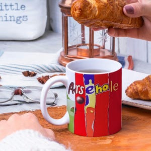 May include: White ceramic mug with a red, yellow, and blue design that says "Arsehole". The mug is filled with a brown liquid and is sitting on a wooden table. There are two croissants on a white plate next to the mug.