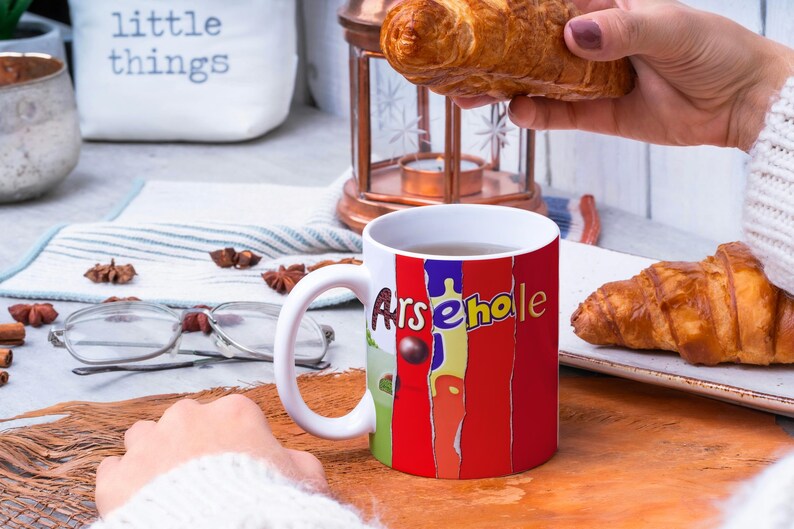 May include: White ceramic mug with a red, yellow, and blue design that says "Arsehole". The mug is filled with a brown liquid and is sitting on a wooden table. There are two croissants on a white plate next to the mug.
