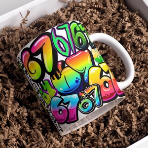 Op de afbeelding: Witte keramische mok met een kleurrijk graffiti-achtig ontwerp met het nummer 676. De nummers zijn in verschillende heldere kleuren, waaronder roze, geel, groen en blauw. De mok heeft een wit handvat en staat in een doos met bruin verpakkingsmateriaal.
