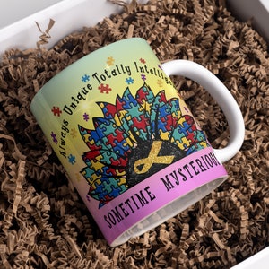Puede incluir: Una taza de cer&aacute;mica blanca con un dise&ntilde;o colorido que presenta un girasol hecho de piezas de rompecabezas. El texto "Always Unique Totally Intelligent Sometime Mysterious" est&aacute; impreso en la taza.