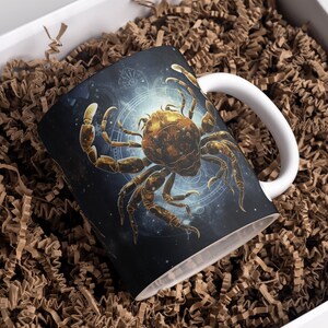 May include: A white ceramic mug with a black and gold design featuring a stylized crab with a celestial background. The mug has a handle on the right side.